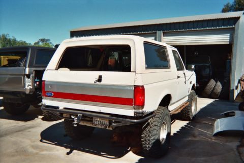 1990 Ford Bronco - Rear three-quarter view of a white lifted Ford Bronco on mud tires parked outside a metal garage with a spare tire nearby.