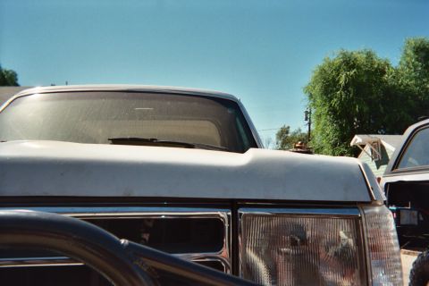 1990 Ford Bronco - Close-up of a vehicle's windshield, cowl and rectangular headlight under a clear blue sky with trees behind.