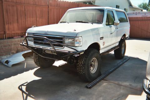 1990 Ford Bronco - white lifted Ford Bronco on mud tires with a black tube front bumper sits on a concrete driveway beside a wood fence.