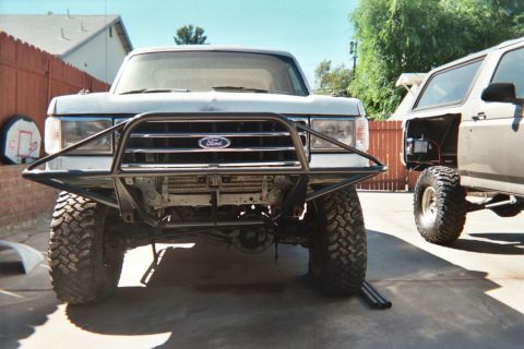 1990 Ford Bronco - Front view of a lifted Ford on mud tires with a black tubular grille guard bearing a Ford emblem, parked by a fence.