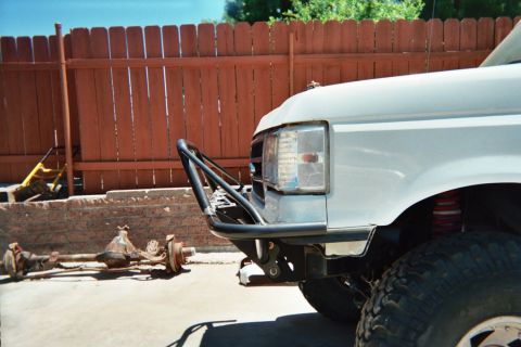 1990 Ford Bronco - Front corner of a white lifted Ford with a black tube bumper; a bare axle assembly lies on the ground by a wood fence.