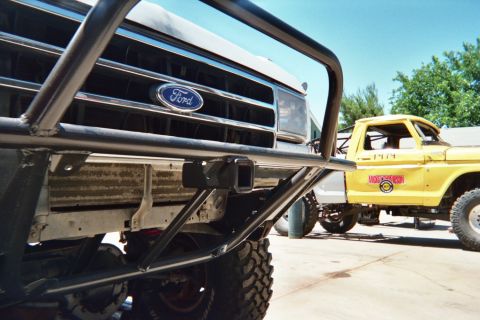 1990 Ford Bronco - Close-up of a Ford grille emblem framed by a black tube bumper, with a yellow caged off-road truck in the background.