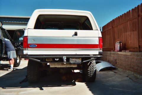 1990 Ford Bronco - Rear view of a white-and-red lifted Ford SUV on mud tires parked on a driveway beside a wood fence and brick wall.