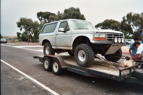 1990 Ford Bronco - white lifted Ford Bronco on mud tires sits loaded on a tandem-axle flatbed trailer in a parking area with people nearby.