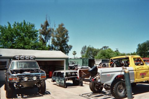 1990 Ford Bronco - Several off-road vehicles, including a yellow caged truck and a light-bar-equipped rig, sit parked at a residential garage.