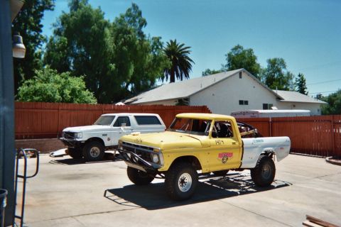 1990 Ford Bronco - yellow lifted vintage Ford pickup with a tube bumper sits beside a white pickup in a fenced backyard driveway.