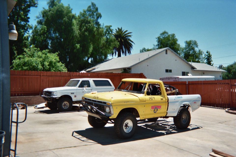 Yellow lifted vintage Ford pickup and white pickup parked in a backyard driveway