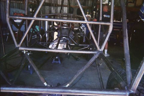 1990 Ford Bronco - bare welded tubular off-road chassis under construction sits in a dim garage with people working in the background.