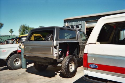 1990 Ford Bronco - gray cut-down off-road SUV with exposed coilover shocks and a roof light bar is parked next to a white Ford Bronco.