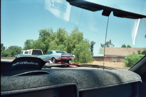 1990 Ford Bronco - Seen through a windshield, a light-blue classic muscle car rides on an open trailer along a highway, with a hat on the dash.