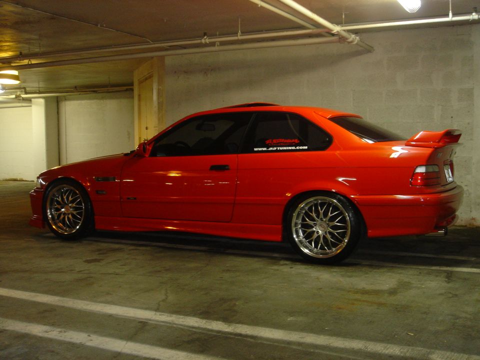 Red BMW E36 M3 coupe side view with mesh wheels and rear wing in a parking garage