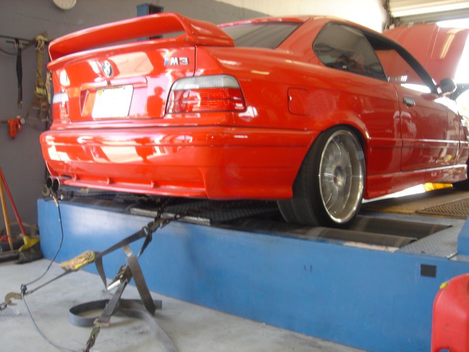 Red BMW M3 coupe with rear wing strapped to a blue dyno, rear wheels on rollers, hood open