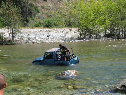 1985 Ford Bronco - A person stands in a blue off-road vehicle navigating a rocky riverbed.
