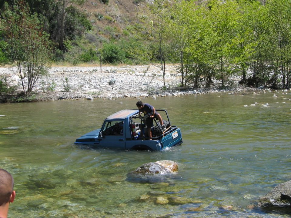 Blue open-top SUV stuck in a shallow rocky river with a man standing on the back