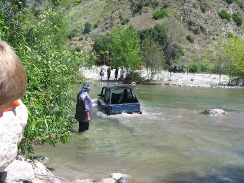1985 Ford Bronco - A blue SUV navigates a shallow river crossing, with several people observing from the bank.