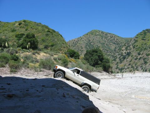 1985 Ford Bronco - Pickup truck navigates a steep dirt embankment in a , hilly landscape under a clear blue sky.
