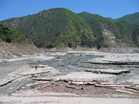 1985 Ford Bronco - A desolate, dry riverbed landscape with debris scattered across the cracked earth, set against a backdrop of green, tree-covered mountains.