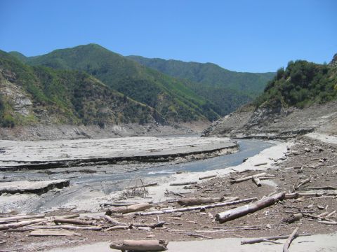 1985 Ford Bronco - A dry riverbed filled with debris and logs, with mountains in the background under a clear blue sky.
