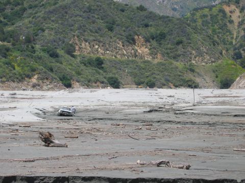 1985 Ford Bronco - A white SUV is partially submerged in mud and debris, surrounded by a desolate, hilly terrain.
