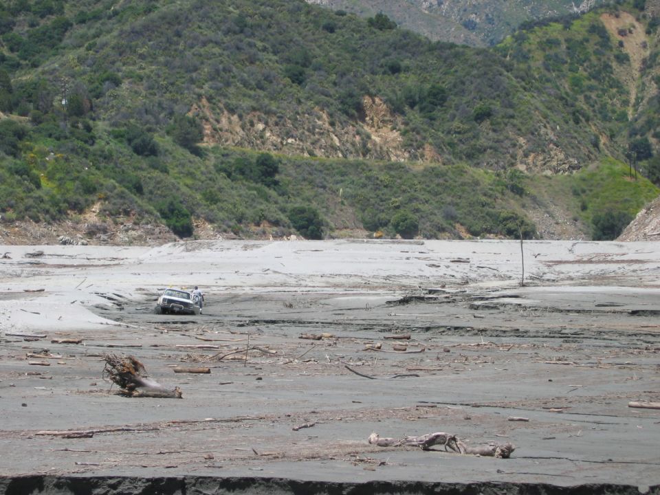 Light-colored SUV stuck in gray mud flats with people beside it, mountains behind.