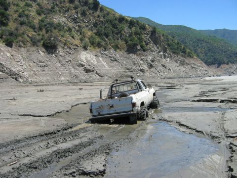 1985 Ford Bronco - A white pickup truck is stuck in a muddy, dry riverbed with steep, vegetated hills in the background.