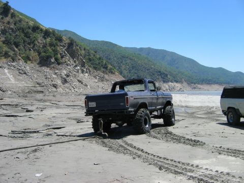 1985 Ford Bronco - This lifted Ford Bronco is driving on a sandy beach near a body of water with mountains in the distance.