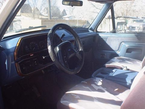 1988 Ford Bronco - Interior of an older Ford truck shot from the driver's door, showing a blue dash and door panels, woodgrain instrument bezel, column shifte…