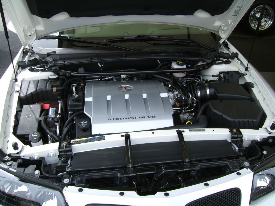 White car engine bay with a silver Northstar V8 engine cover. A SuperMotors.net logo is visible at the bottom right