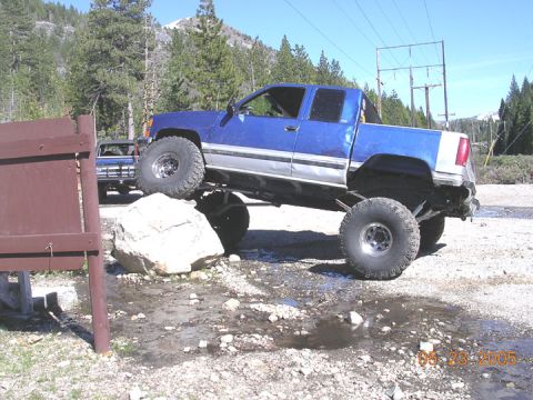 1994 Ford Bronco - A lifted blue pickup truck navigates over a large rock in a rocky, outdoor area.