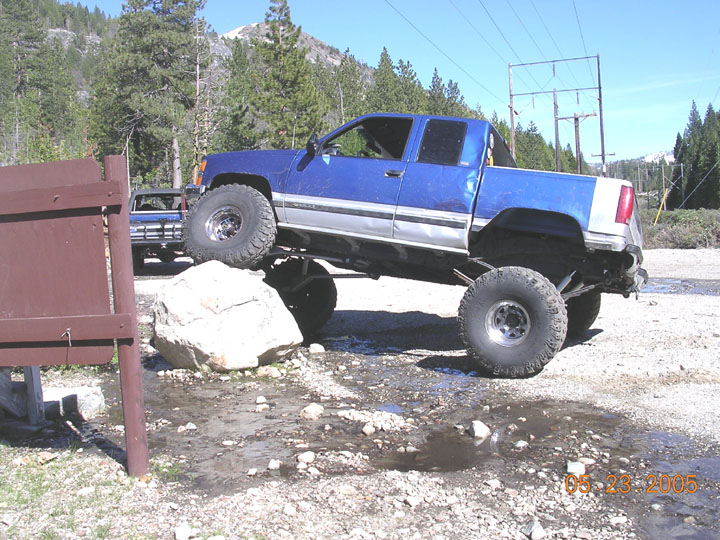 Lifted blue and silver extended-cab pickup flexed over a boulder on a mountain trail
