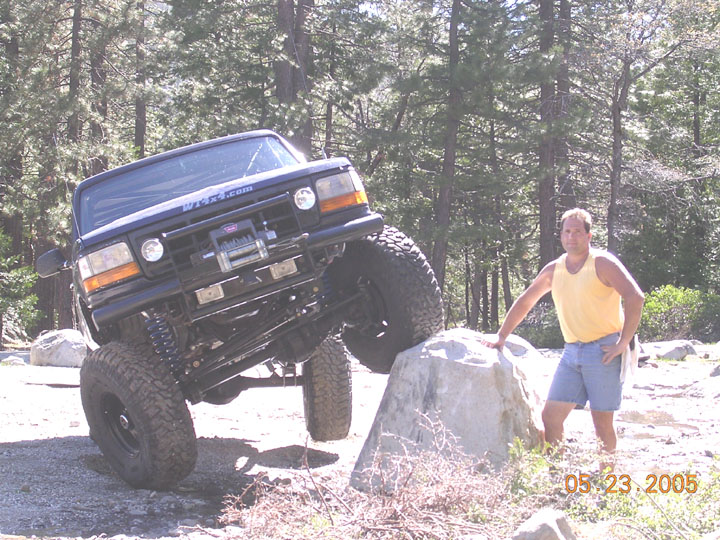 Lifted dark green Ford Bronco with winch flexing front axle over a boulder in pine forest