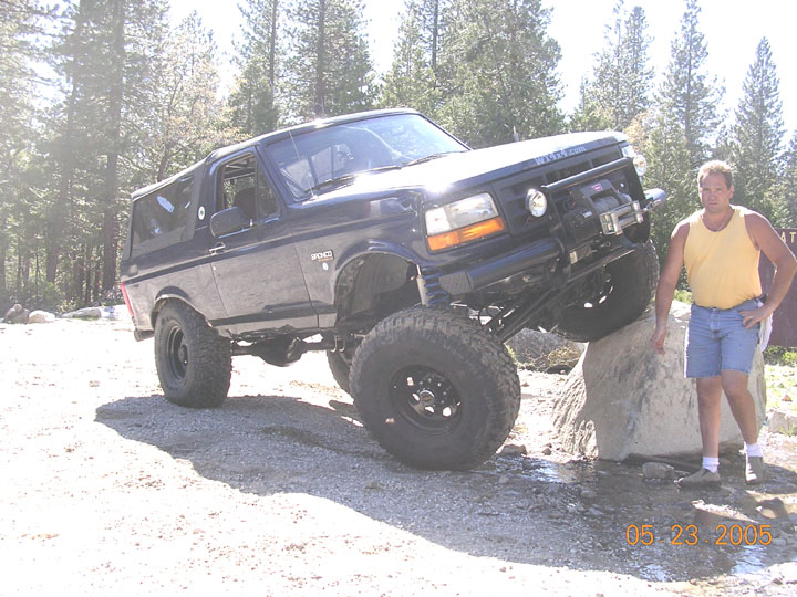 Black lifted Ford Bronco with winch bumper flexing over a rock on a pine-lined trail