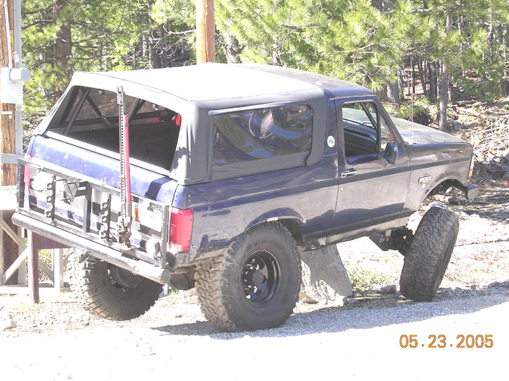 Dark blue Ford Bronco with soft top flexing over a boulder, Hi-Lift jack on the rear tube bumper