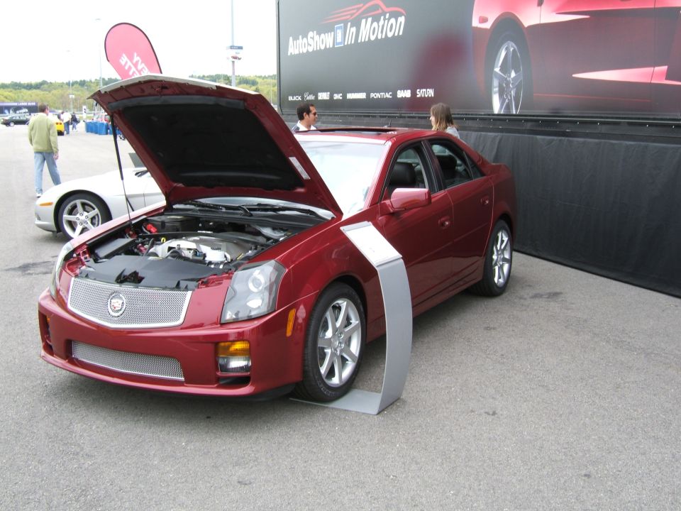 Red Cadillac sedan, hood open, showing engine. Silver trim piece in front of front wheel. Other cars and a banner in background