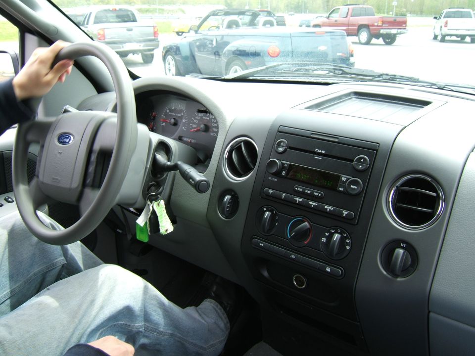 Interior view of a pickup truck dashboard with a hand on the steering wheel; other trucks visible outside