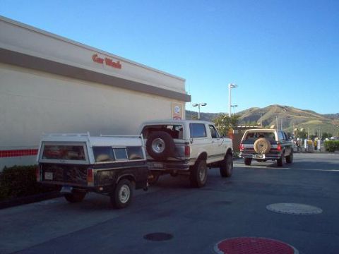 1988 Ford Bronco - Two Ford Broncos and a pickup truck are parked outside a car wash on a sunny day with hills in the background.