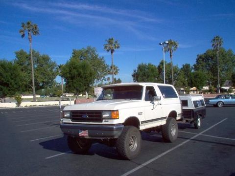 1988 Ford Bronco - A white Ford Bronco is parked in a lot, towing a trailer under a clear blue sky.