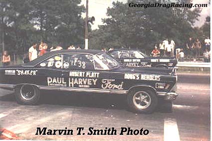 1967 Ford Fairlane - A black 1966 Ford Fairlane drag car with extensive lettering races down the strip, strip next to another vehicle.