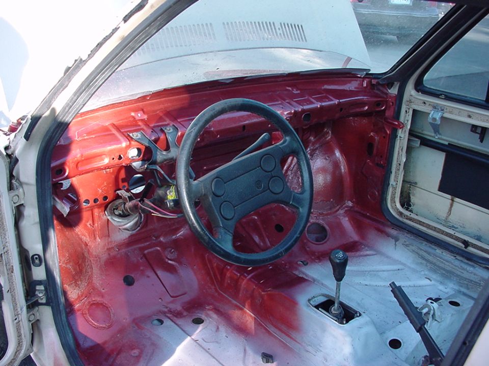Car interior, stripped down with red primer on the dash and floor. Features a black steering wheel and manual shifter