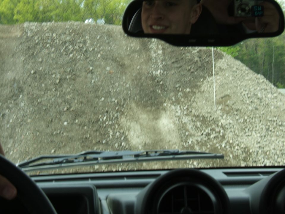 driver's perspective looking out windshield at a large pile of gravel, with a person visible in the rearview mirror