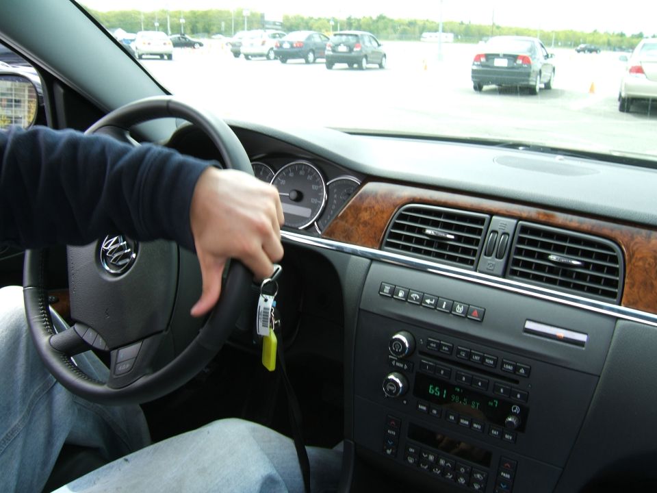 Driver's hand on steering wheel, dashboard with wood trim, air vents, and radio visible. Other cars in parking lot