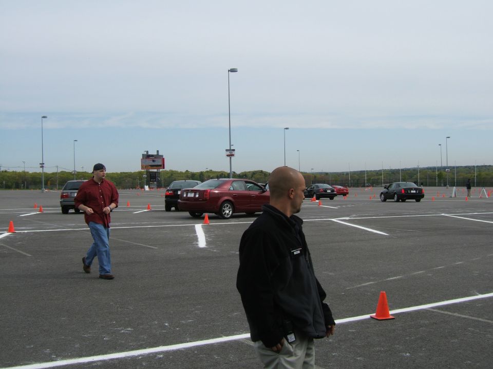 Parking lot with several cars, orange cones, and people. A scoreboard is visible in the distance