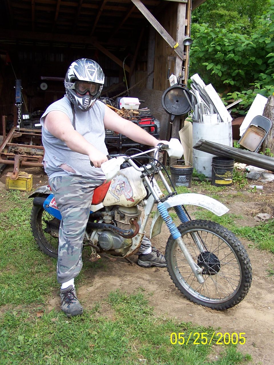 Rider in helmet on worn white and red dirt bike outside a cluttered barn