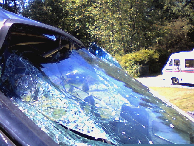 Shattered, collapsed windshield on a dark SUV with crushed roof; white RV in grassy background