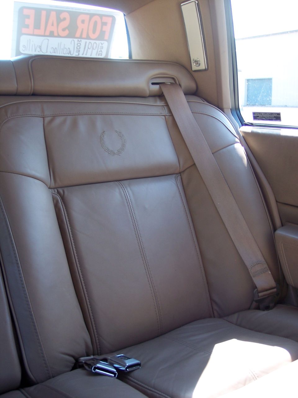 Tan leather rear seat of a 1991 Cadillac DeVille with embroidered wreath emblem and for-sale sign in window
