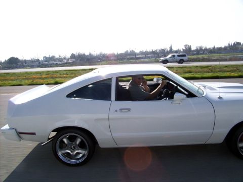 1978 Ford Mustang II - A white 1975 Ford Mustang II coupe is driven by a man on a highway, with another vehicle in the background.