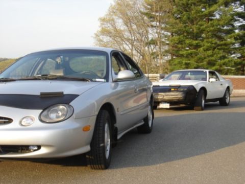 1999 Ford Taurus - A silver 1999 Ford Taurus is parked on the street, with another car visible in the background.