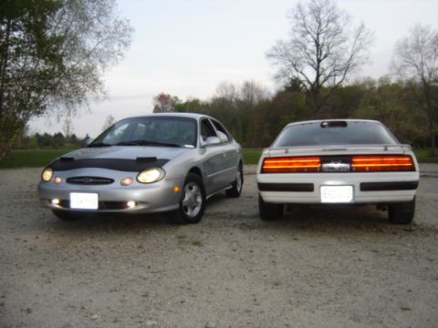 1999 Ford Taurus - Two sedans are parked side-by-side on a gravel surface with trees in the background.