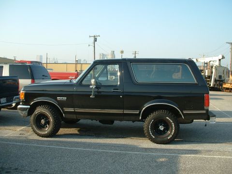 1989 Ford Bronco - A black 1989 Ford Bronco is parked in a lot with other vehicles and industrial equipment visible.