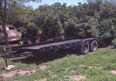 1995 Ford Bronco - A dark tandem-axle flatbed car-hauler trailer with a wood deck and steel drive-over fenders, parked on grass in front of trees. An older ro…